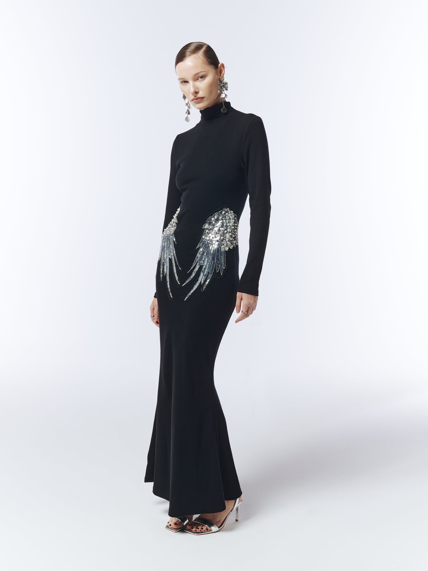 Model wearing long sleeve black evening gown with silver sequin wing details at hips.