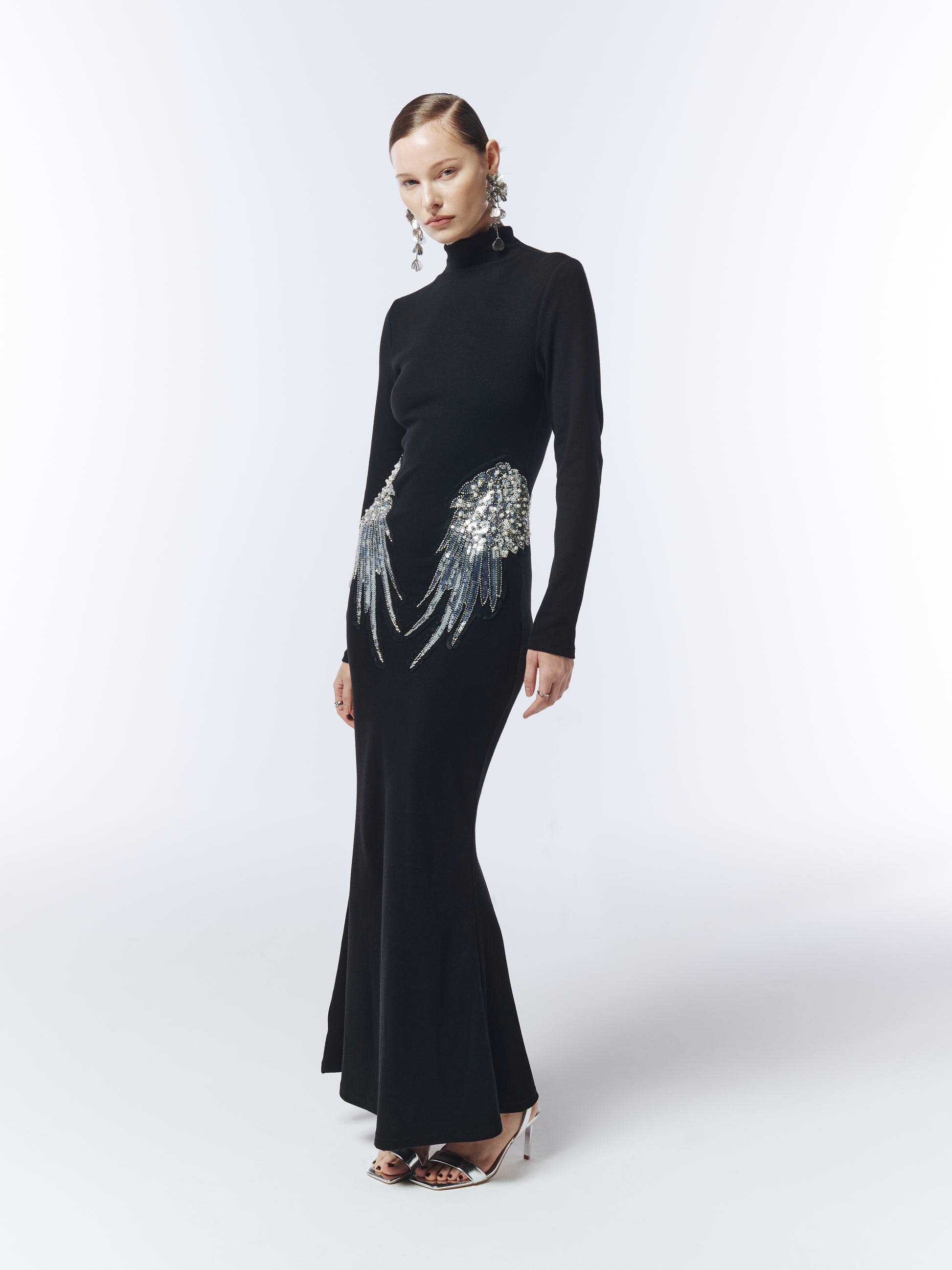 Model wearing long sleeve black evening gown with silver sequin wing details at hips.