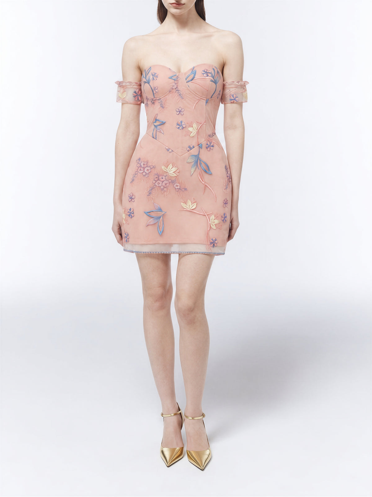 Model wearing a SOUR FIGS blush-pink off-the-shoulder mini dress with a sweetheart neckline, fitted bodice and delicate blue, pink and cream floral embroidery, styled with red ankle-strap heels.