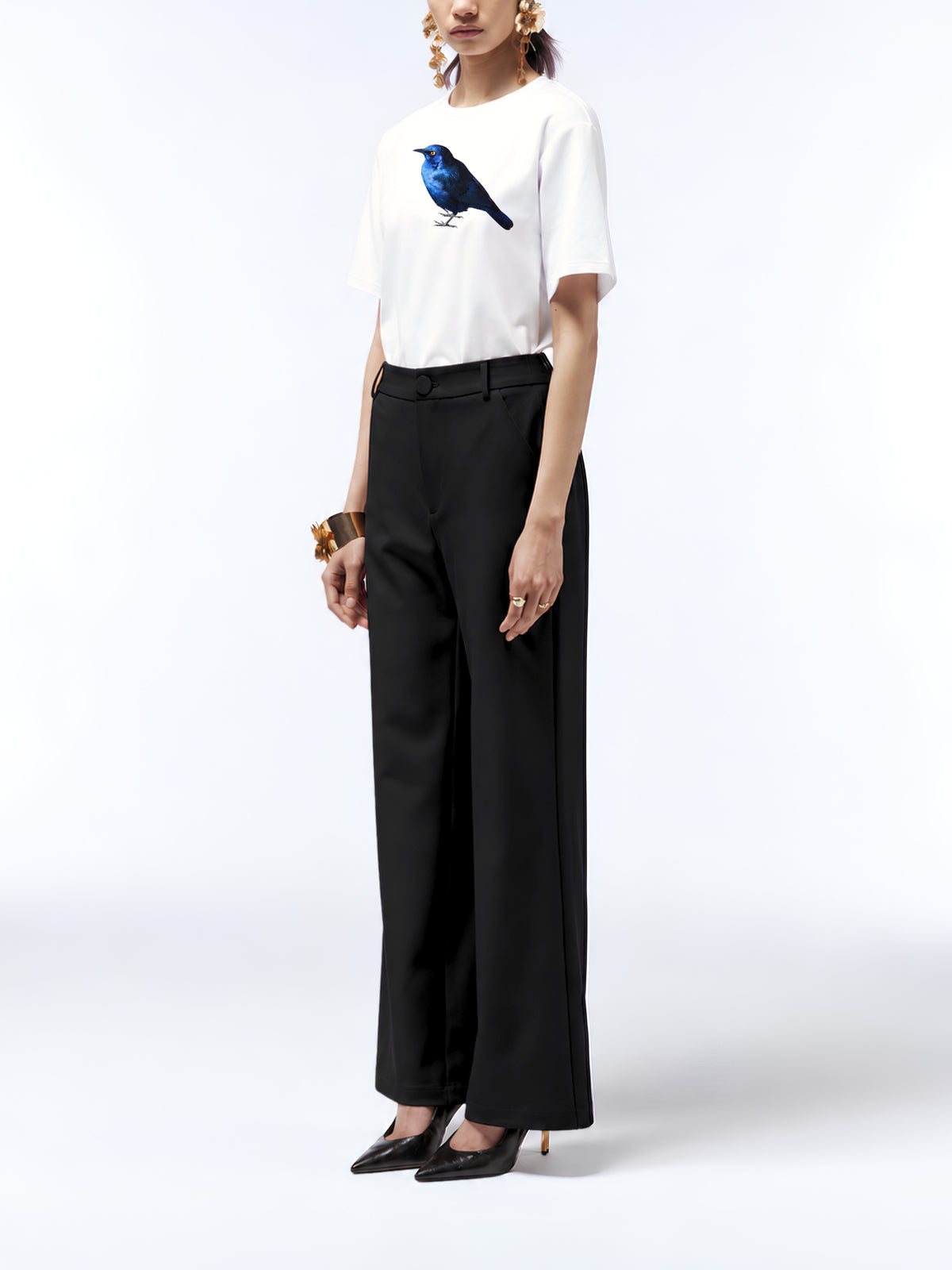 Model wearing the white starling print T-shirt untucked with black trousers, pointed heels and gold jewellery.