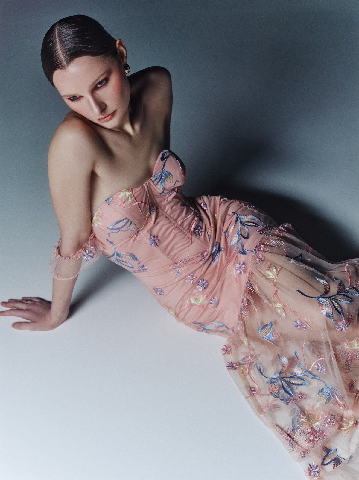 Blush pink floral-embroidered bustier maxi dress, seated three-quarter model view showing the pointed waist seam and sheer skirt.