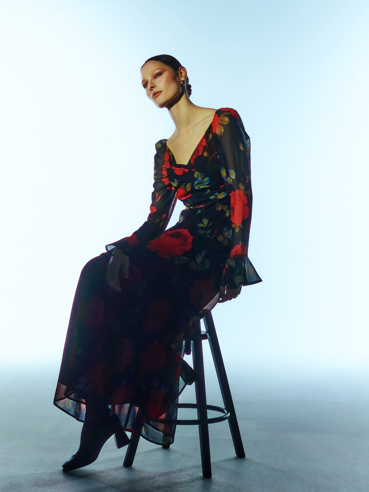 Seated three-quarter model view of the SOUR FIGS Floral Long-Sleeve Maxi Dress in Midnight Rose Black, with red roses, a ruched V-neck bodice and sheer sleeves with flared cuffs.